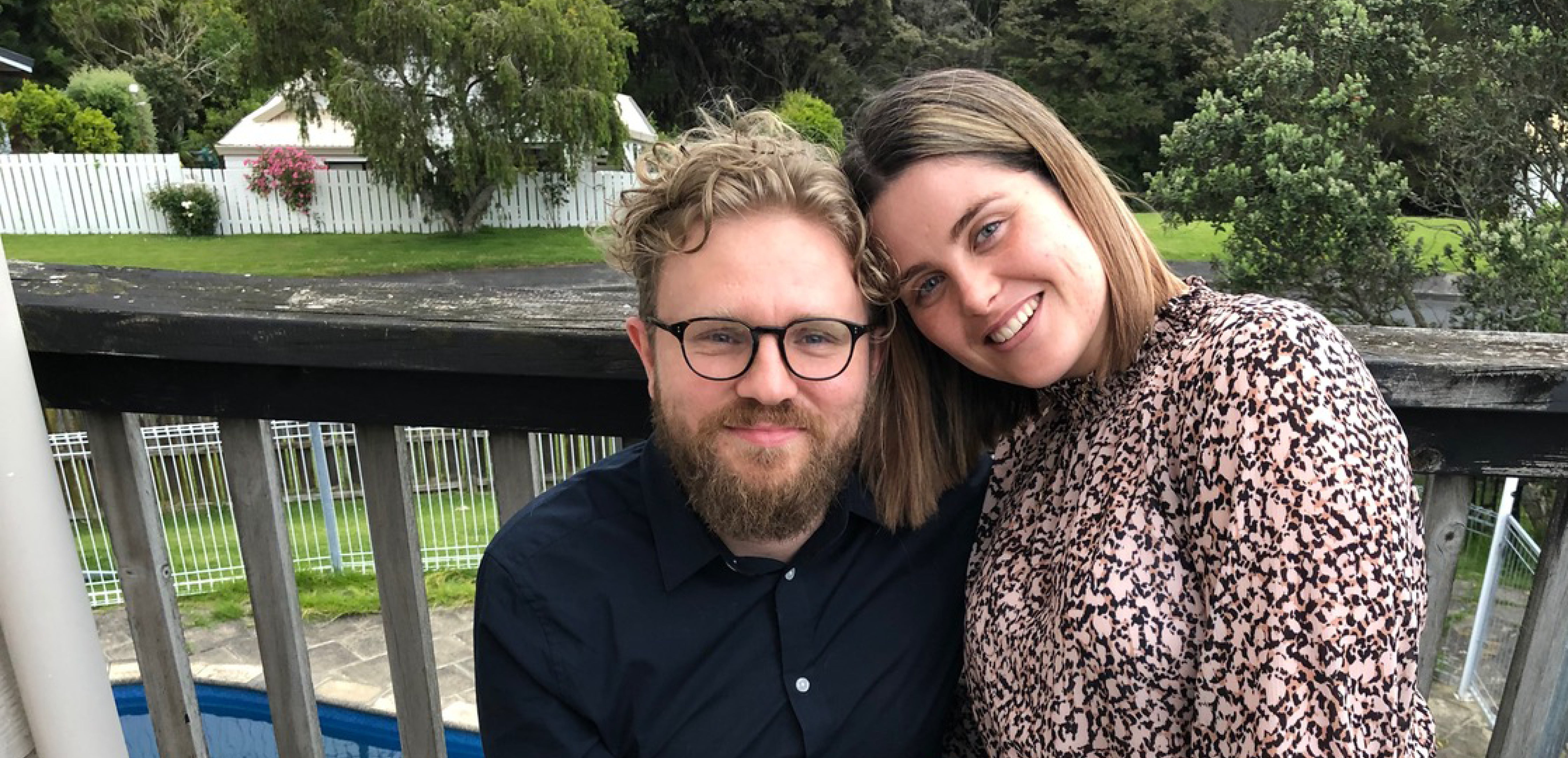 Alt text: Couple sitting and embracing on a backyard balcony with the woman’s heads resting against her partner.