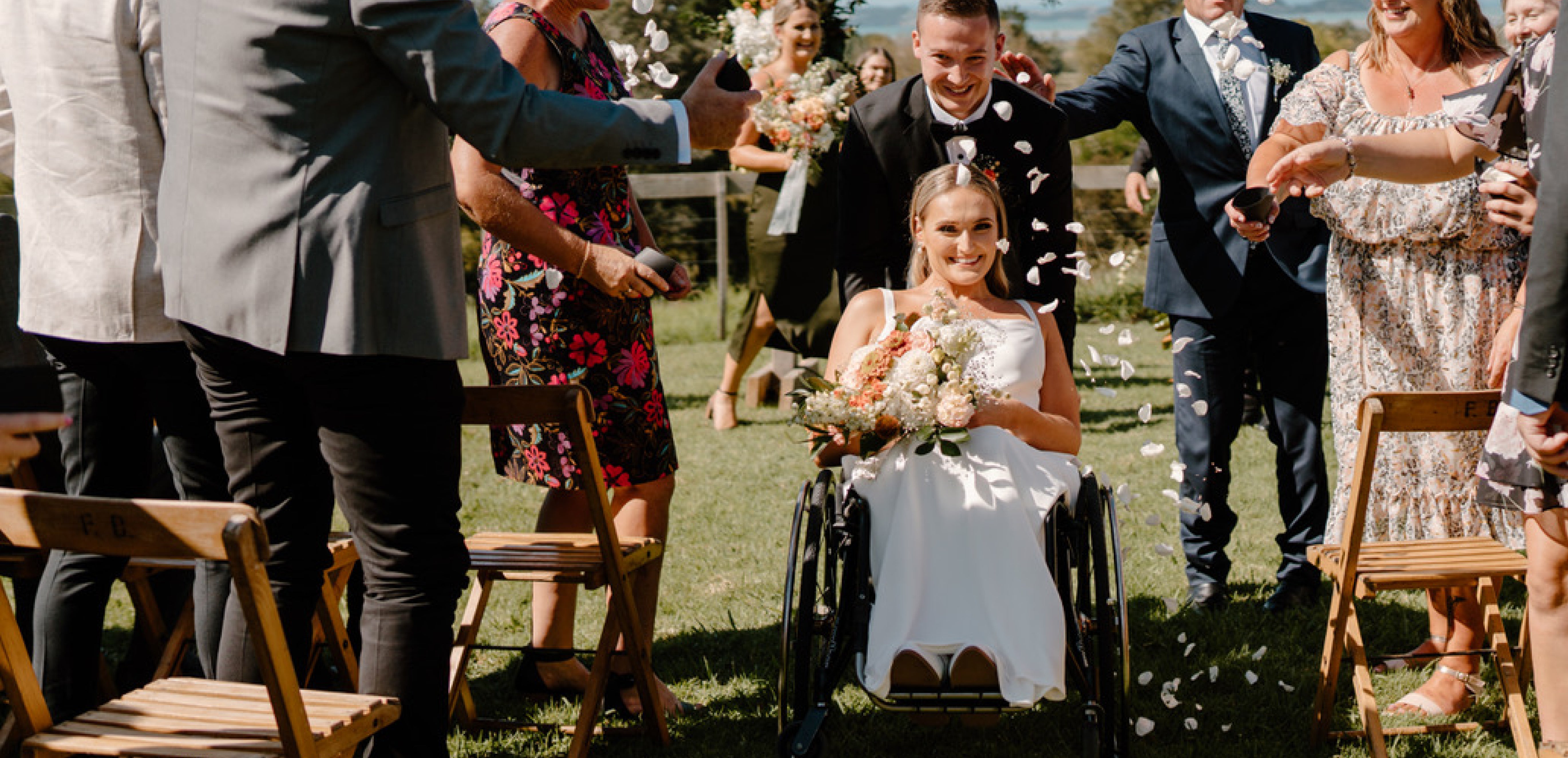 Alt text: Bride in wheelchair being pushed down the aisle by husband at outside wedding & with flower petals being tossed by guests.