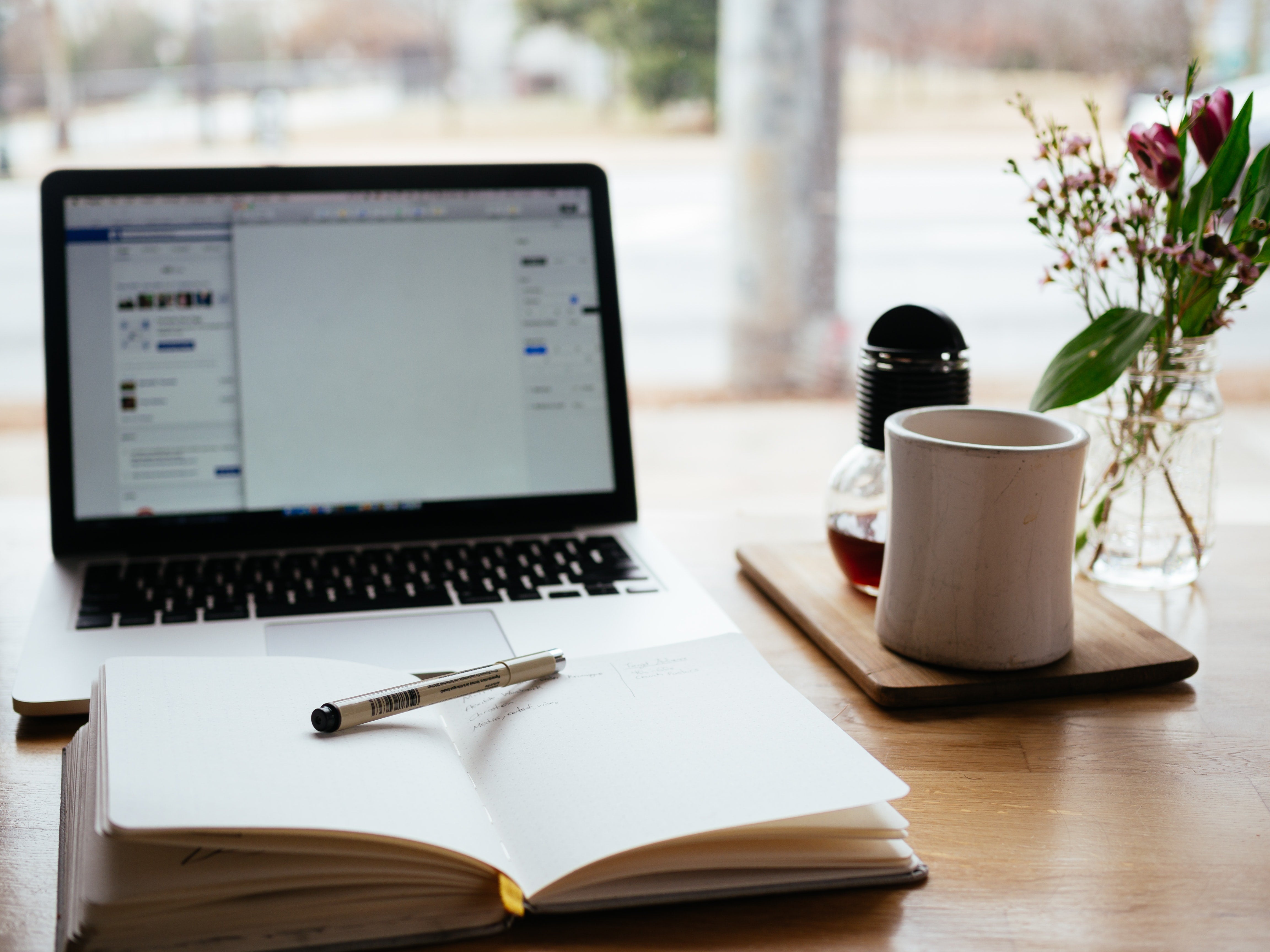 Alt text: A workstation on wooden desk with open diary, laptop, and coffee with a blurred view of outside.